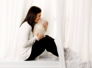 Mother gently holding and kissing her newborn baby for baby photo shoot, wrapped in a soft knitted blanket, sitting by a window for professional newborn photography.